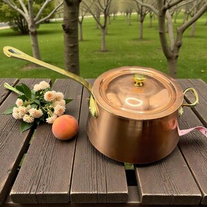 May include: A copper saucepan with a lid and a long brass handle. The pot is sitting on a wooden table with a peach and a small bouquet of flowers. The background shows a green lawn and trees.