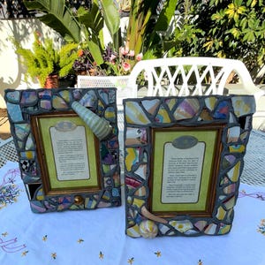 Puede incluir: Dos marcos de fotos rectangulares con un borde de mosaico hecho de fragmentos de cer&aacute;mica de colores incrustados en lechada gris. Cada marco incluye un borde interior dorado y una tarjeta impresa con texto descriptivo.