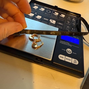 May include: Gold hoop earrings on a digital scale, with a measuring tape showing a length of approximately 1.5 inches. The scale displays a weight of 0.01g. The scale has a black surface with white text and buttons.