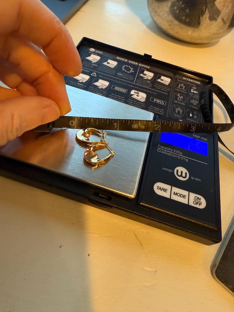 May include: Gold hoop earrings on a digital scale, with a measuring tape showing a length of approximately 1.5 inches. The scale displays a weight of 0.01g. The scale has a black surface with white text and buttons.