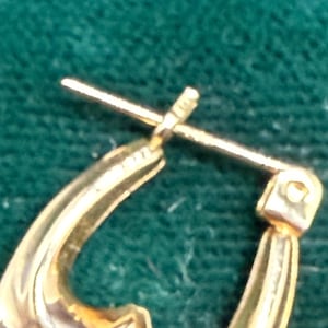 May include: Close-up of a gold-colored hoop earring. The earring has a curved design with a post and clasp closure. The metal has a polished finish, reflecting light. The background is a dark green, textured surface.