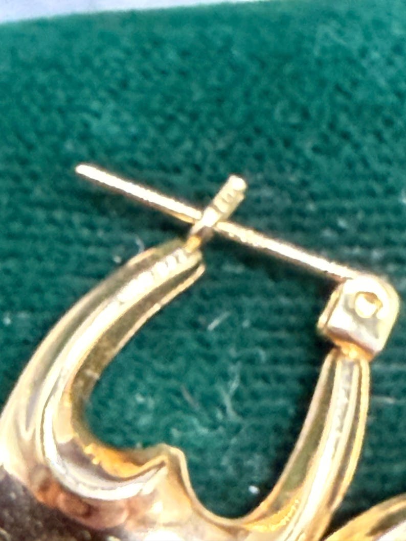 May include: Close-up of a gold-colored hoop earring. The earring has a curved design with a post and clasp closure. The metal has a polished finish, reflecting light. The background is a dark green, textured surface.
