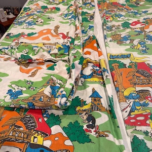 May include: A white bed sheet and pillowcase set featuring a colorful Smurf cartoon print. The pattern displays blue Smurf characters, orange and red mushroom houses, green grass, and the text SMURF in yellow letters on a blue banner background.