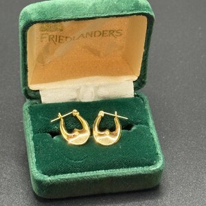 May include: A pair of gold hoop earrings with a heart-shaped design, displayed in an open, green velvet jewelry box. The box has a cream-colored interior and the text "FRIEDLANDERS" is visible on the inside of the lid.