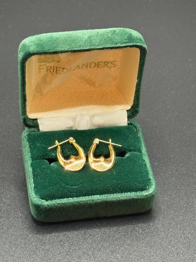 May include: A pair of gold hoop earrings with a heart-shaped design, displayed in an open, green velvet jewelry box. The box has a cream-colored interior and the text "FRIEDLANDERS" is visible on the inside of the lid.