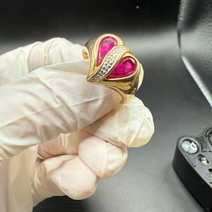 May include: A gold ring with two heart-shaped ruby gemstones. The ring has a band of small diamonds between the ruby hearts. The ring is set against a dark background.