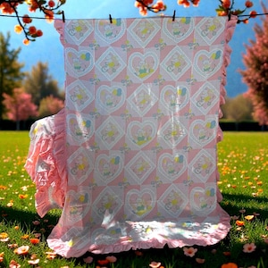 May include: A pink baby blanket with a pattern of hearts, bunnies, and bicycles, hanging on a washing line. The blanket has a frilled edge and is displayed outdoors on a sunny day.