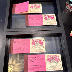 May include: Two black framed shadow boxes displaying vintage Disneyland tickets. The tickets are pink and tan with the Disneyland logo and text. The frames are rectangular and have a dark finish. The tickets are from the Walt Disney Story.