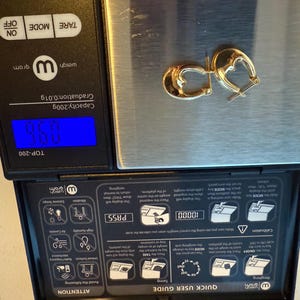 May include: A pair of gold-colored hoop earrings resting on a digital scale. The scale displays a weight of 9.60 grams. The earrings have a polished finish and a classic design. The scale is black with a blue digital display.