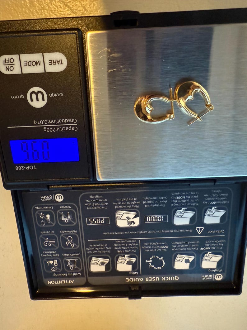 May include: A pair of gold-colored hoop earrings resting on a digital scale. The scale displays a weight of 9.60 grams. The earrings have a polished finish and a classic design. The scale is black with a blue digital display.