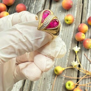 May include: A gold ring with two heart-shaped ruby stones and a row of small diamonds. The ring is held by a gloved hand, with peaches and small apples scattered on a wooden surface.