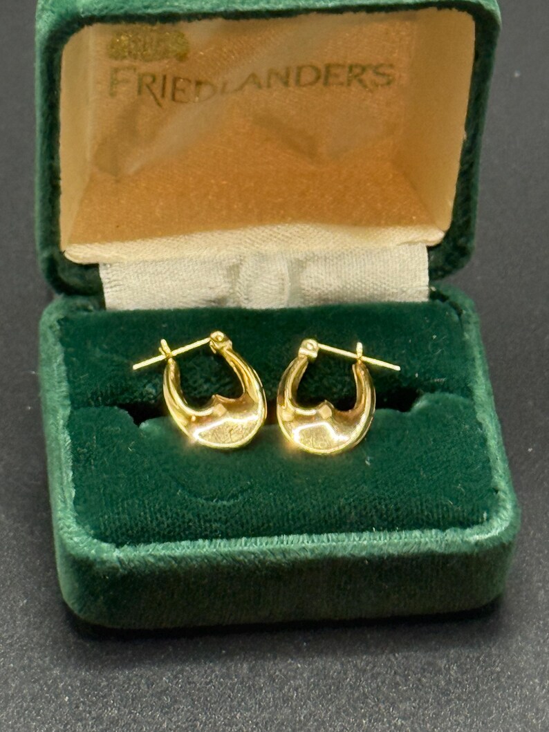 May include: A pair of gold hoop earrings displayed in a vintage green velvet jewelry box. The earrings have a polished finish and a classic design. The box has a gold satin interior and the name "FRIEDLANDERS" printed on the inside.