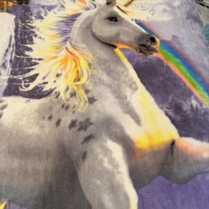 Può includere: Una coperta in peluche con un motivo a unicorno. L'immagine presenta un unicorno bianco con un corno dorato e un arcobaleno. Lo sfondo presenta un castello e un cielo stellato nei toni del viola e del blu.