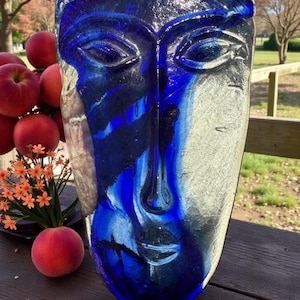 May include: A blue and clear glass face sculpture with a serene expression. The sculpture has a long nose, closed lips, and detailed eyes. The glass is a mix of deep blue and clear, creating a striking contrast. The sculpture is displayed on a wooden surface.