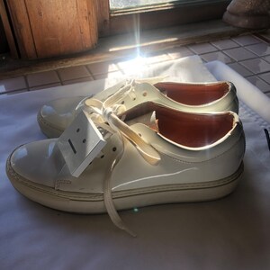 May include: A pair of white leather low-top trainers with white laces. The trainers have a white rubber sole and a small white tag with a black line on it.