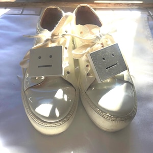 May include: A pair of white leather low-top trainers with white laces and a square face graphic on each shoe. The graphic is a simple black line drawing of a face with a straight line for a mouth.