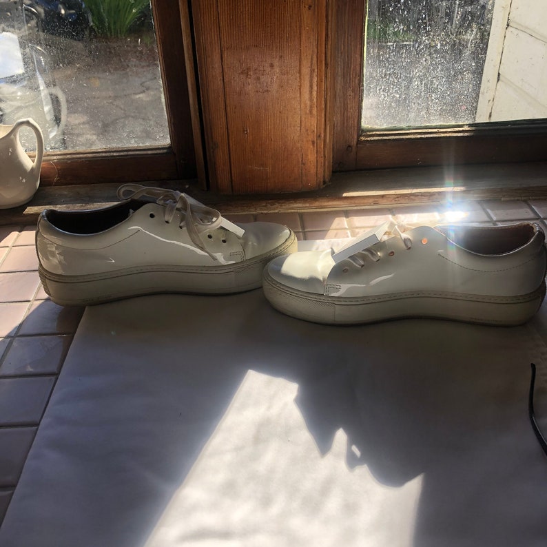 May include: A pair of white patent leather platform trainers with white laces. The trainers are on a white tiled floor.