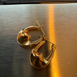 May include: A pair of gold-colored hoop earrings. The earrings have a smooth, polished surface and a teardrop shape. The earrings are displayed on a metallic surface.