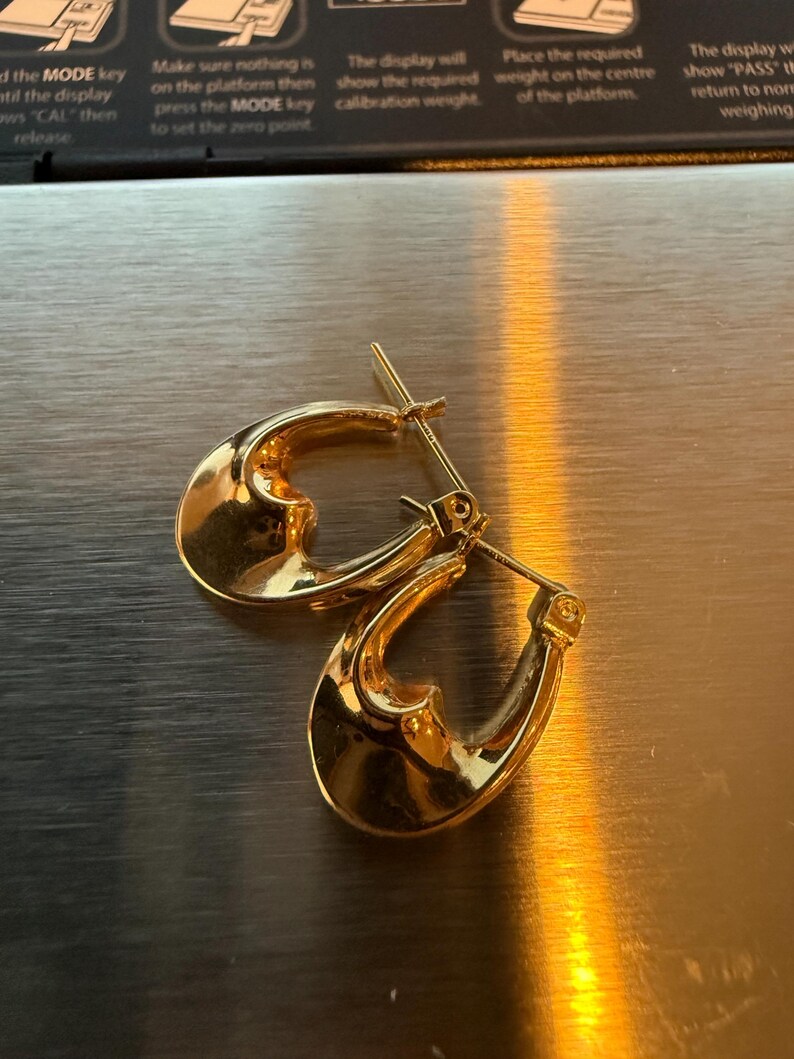 May include: A pair of gold-colored hoop earrings. The earrings have a smooth, polished surface and a teardrop shape. The earrings are displayed on a metallic surface.