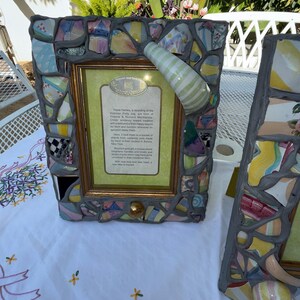 Puede incluir: Un marco de fotos rectangular con un borde de mosaico hecho de fragmentos de cer&aacute;mica de colores fijados en lechada gris. Un asa de cer&aacute;mica a rayas verdes y blancas est&aacute; unida a la esquina superior derecha. El marco contiene una tarjeta con texto.