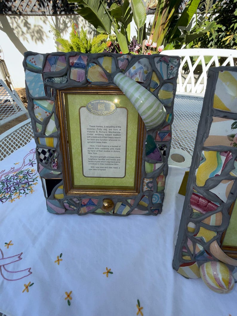 Puede incluir: Un marco de fotos rectangular con un borde de mosaico hecho de fragmentos de cer&aacute;mica de colores fijados en lechada gris. Un asa de cer&aacute;mica a rayas verdes y blancas est&aacute; unida a la esquina superior derecha. El marco contiene una tarjeta con texto.