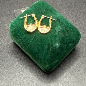 May include: A pair of gold-colored hoop earrings with a decorative design, displayed on a dark green velvet jewelry box. The earrings have a curved shape with a detailed interior. The background is a dark, textured surface.