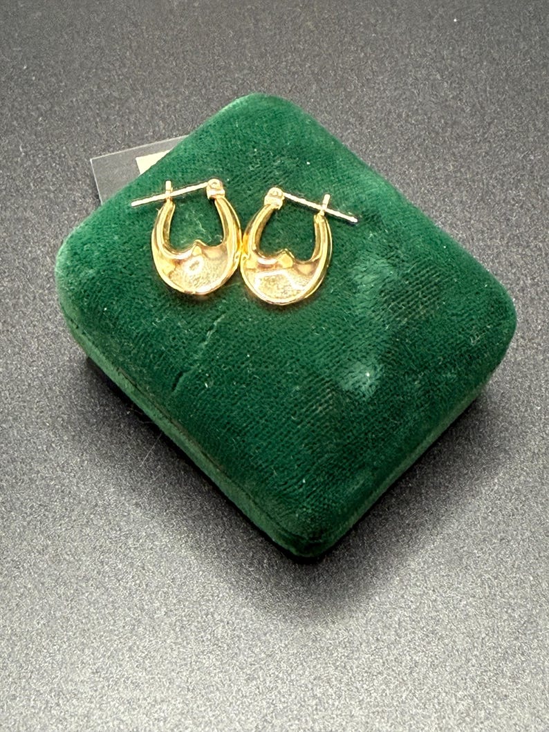 May include: A pair of gold-colored hoop earrings with a decorative design, displayed on a dark green velvet jewelry box. The earrings have a curved shape with a detailed interior. The background is a dark, textured surface.