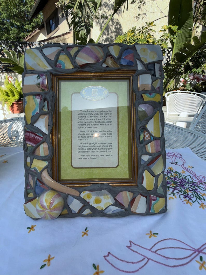 Puede incluir: Un marco de fotos rectangular con un borde de mosaico hecho de fragmentos de cer&aacute;mica multicolor fijados en mortero gris. El marco incluye un borde interior dorado y una inserci&oacute;n central con texto impreso sobre el reciclaje de cer&aacute;mica.