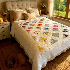 May include: A bed featuring a cream-colored quilt with a multi-colored geometric triangle pattern, topped with two large beige pillows and a smaller floral accent pillow, set against a tufted headboard with a sleeping dog on the floor.