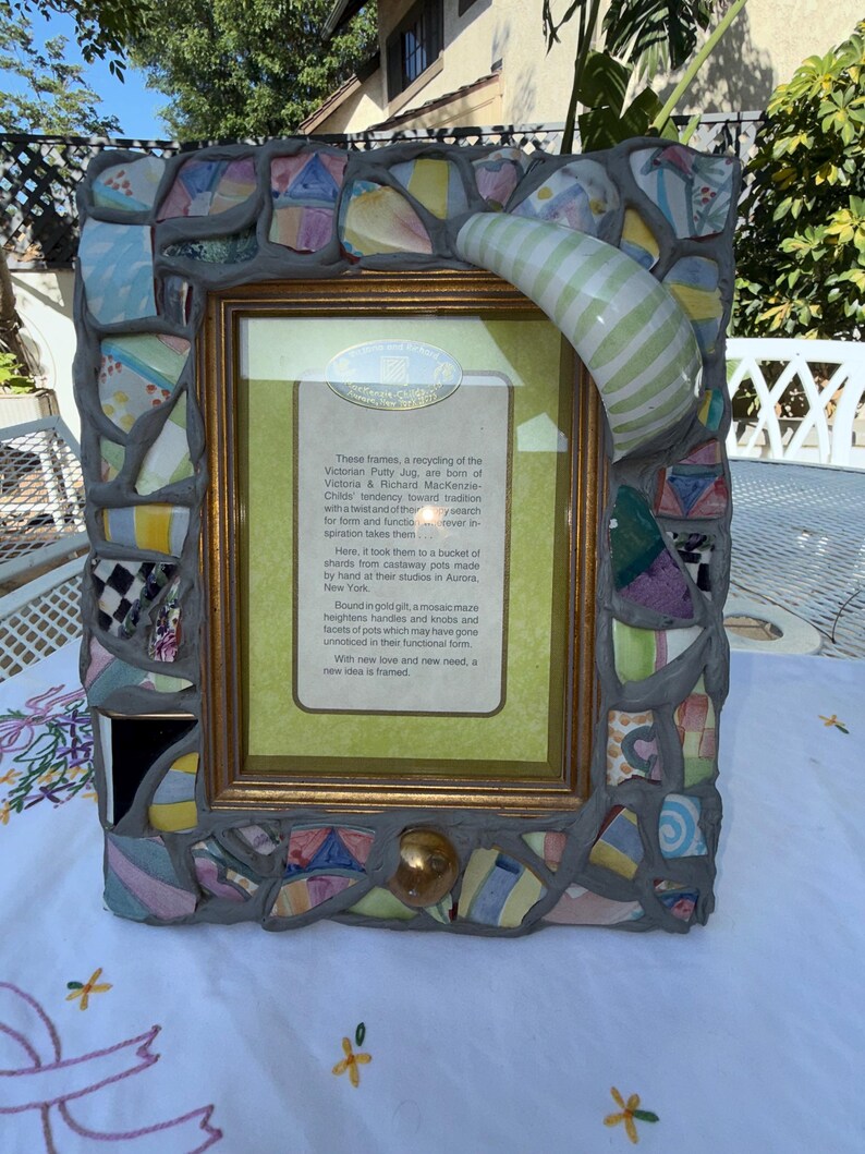 Puede incluir: Un marco de fotos rectangular con un borde de mosaico hecho de fragmentos de cer&aacute;mica multicolor fijados en lechada gris. El marco incluye un asa de cer&aacute;mica a rayas verdes y blancas que sobresale y un pomo dorado. Una tarjeta central contiene texto.