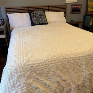 May include: A cream-coloured bedspread with a textured pattern and fringe trim. The bed has white pillows and a black decorative pillow with a white abstract design. The headboard is made of wood with horizontal slats.