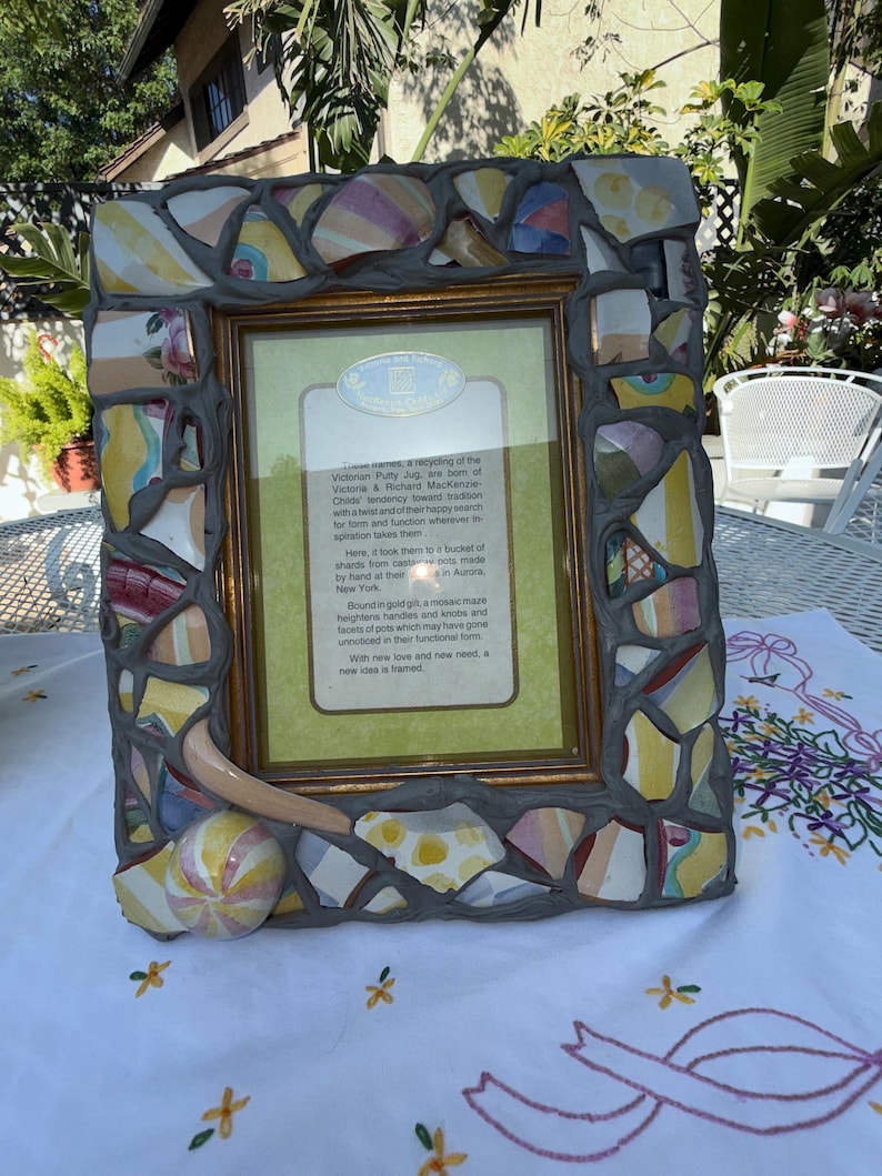 Puede incluir: Un marco de fotos rectangular con un borde de mosaico hecho de fragmentos de cer&aacute;mica coloridos fijados en lechada gris. El marco incluye un asa de cer&aacute;mica que sobresale y un acento esf&eacute;rico. Una tarjeta impresa con texto est&aacute; centrada.
