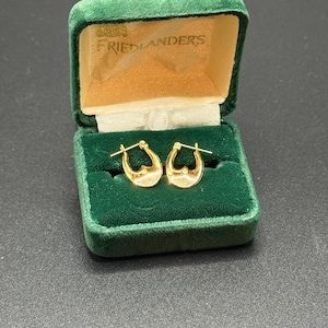 May include: A pair of gold hoop earrings displayed in an open, vintage green velvet jewelry box. The box has a gold satin lining and the name "FRIEDLANDERS" printed on the inside of the lid. The earrings are small, with a textured surface.