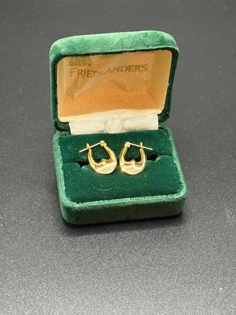 May include: A pair of gold hoop earrings displayed in an open, vintage green velvet jewelry box. The box has a gold satin lining and the name "FRIEDLANDERS" printed on the inside of the lid. The earrings are small, with a textured surface.