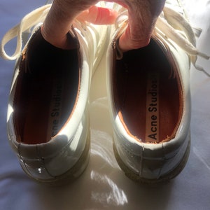 May include: A pair of white leather sneakers with a glossy finish. The inside of the shoes are lined with brown leather and have the text "Acne Studios" printed on the insole.