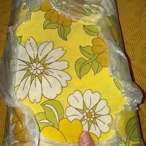 May include: A folded flat sheet packaged in clear plastic, featuring a vibrant yellow background with a large-scale floral pattern in shades of white, light green, and golden yellow. A small white tag labelled FLAT is visible at the top edge.