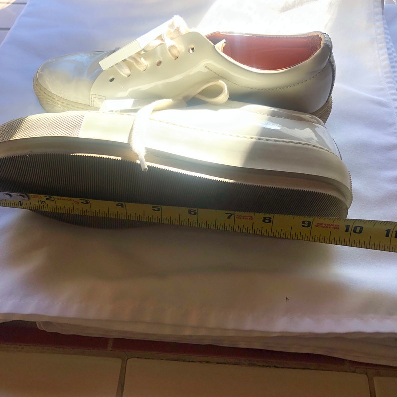 May include: White leather sneakers with a white rubber sole. The sneakers are measured with a tape measure, showing a size of 9 inches.