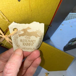 Puede incluir: Una parte posterior de marco de fotos cubierta de tela color amarillo mostaza con una etiqueta de papel beige sujeta por un lazo de rafia. La etiqueta muestra una silueta de conejo, un dise&ntilde;o de corona y el texto THE ROSEMARY RABBIT SARASOTA.FL.