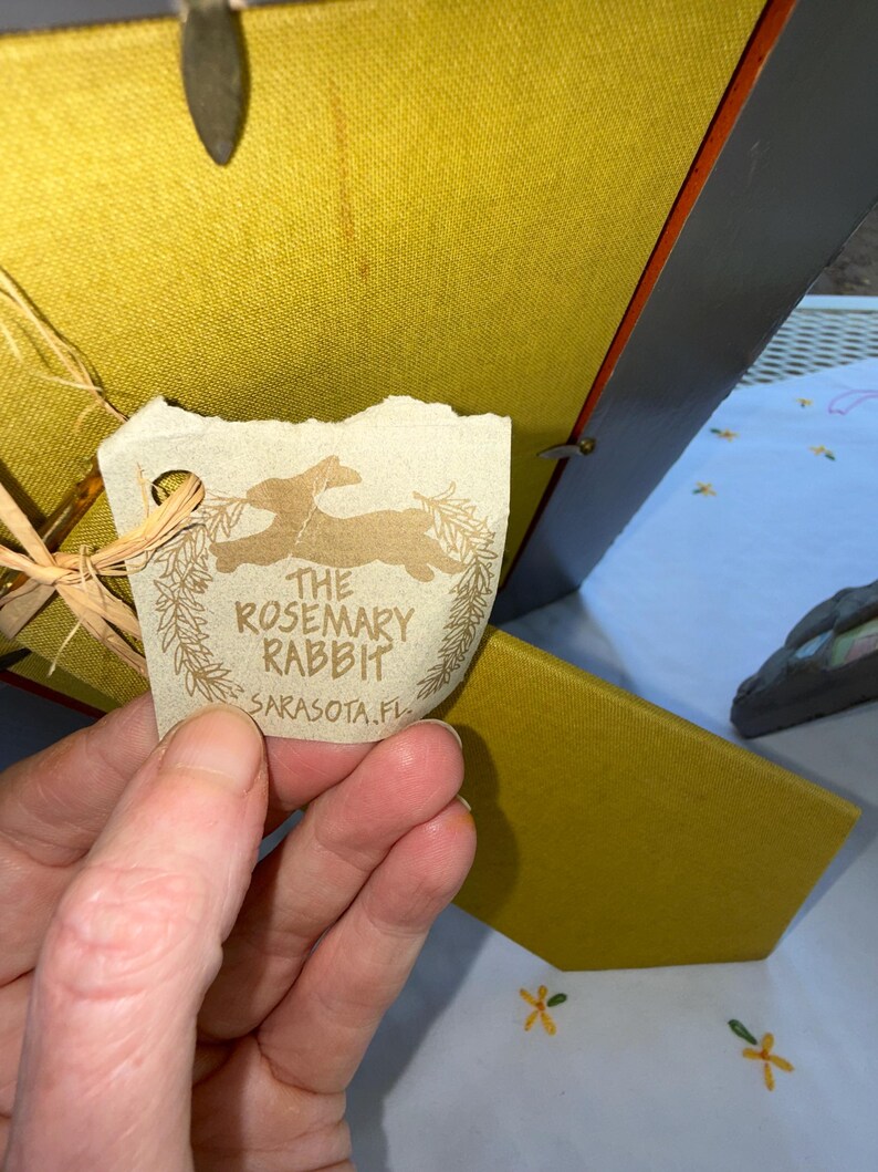 Puede incluir: Una parte posterior de marco de fotos cubierta de tela color amarillo mostaza con una etiqueta de papel beige sujeta por un lazo de rafia. La etiqueta muestra una silueta de conejo, un dise&ntilde;o de corona y el texto THE ROSEMARY RABBIT SARASOTA.FL.
