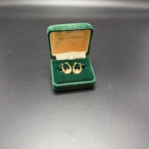 May include: A pair of gold hoop earrings displayed in an open green velvet jewelry box. The box has a gold interior and the name "FRIEDLANDERS" printed on the inside of the lid. The earrings are small and crescent-shaped.