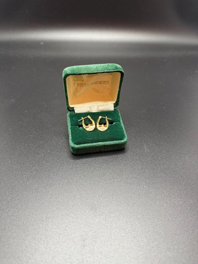 May include: A pair of gold hoop earrings displayed in an open green velvet jewelry box. The box has a gold interior and the name "FRIEDLANDERS" printed on the inside of the lid. The earrings are small and crescent-shaped.