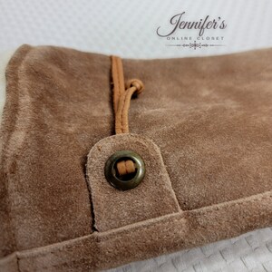 May include: Close-up of a brown suede bag with a brown leather strap and a metal button. The bag has a soft, textured surface.