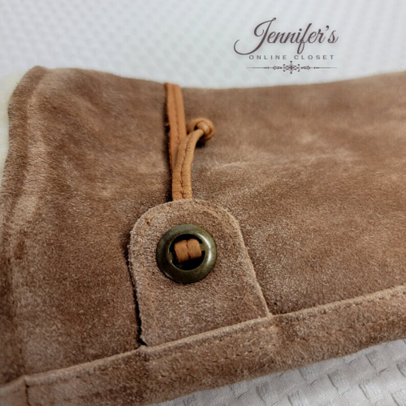 May include: Close-up of a brown suede bag with a brown leather strap and a metal button. The bag has a soft, textured surface.