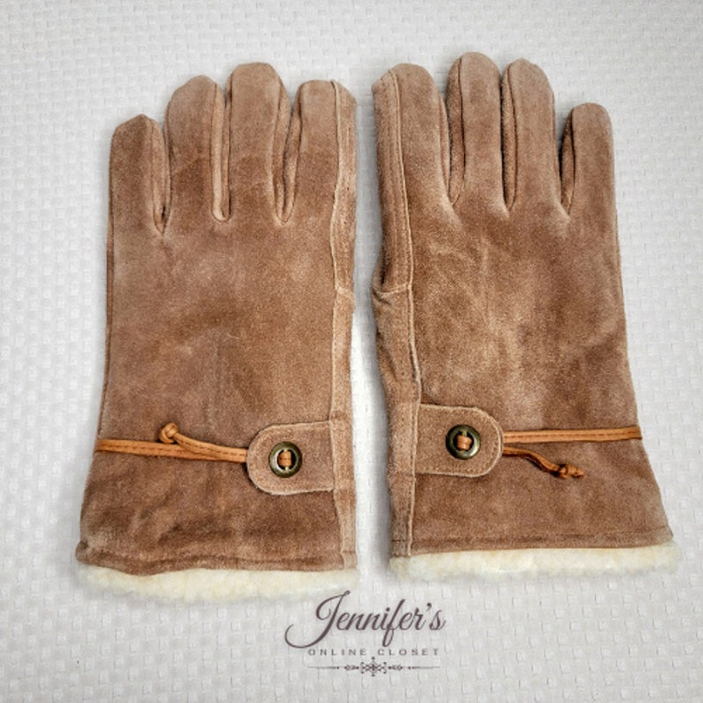 May include: A pair of brown suede gloves with a brown leather strap and a metal button. The gloves have a white fleece lining.