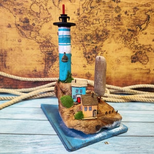 Può includere: Un faro in miniatura con una torre a strisce blu e bianche, una piccola barca a vela e una base in legno. Il faro è circondato da case in miniatura e un piccolo albero. La scena è posizionata su una base in legno con un bordo dipinto di blu.