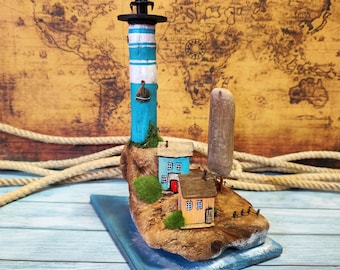 May include: A miniature lighthouse with a blue and white striped tower, a small sailboat, and a wooden base. The lighthouse is surrounded by miniature houses and a small tree. The scene is set on a wooden base with a blue painted edge.