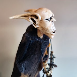 May include: A white, sculpted figure with pointed ears and a stern expression. The figure is wearing a black cloak and has a necklace with a yellow pendant. The figure is mounted on a dark, wooden base.