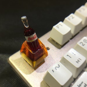May include: A custom mechanical keyboard keycap featuring a miniature dark brown Coca-Cola bottle embedded within a translucent amber-colored resin base. The bottle has a white label with the red Coca-Cola logo and a silver cap, positioned on a keyboard.