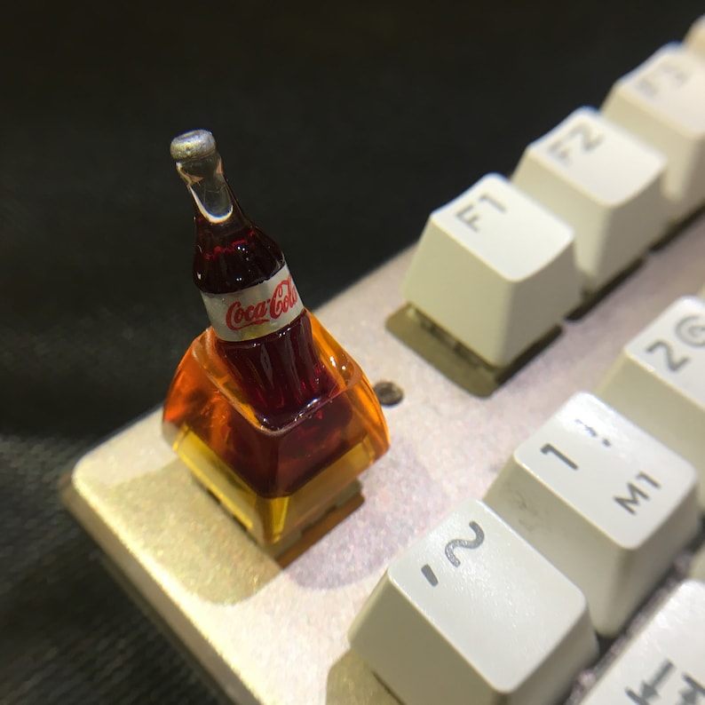 May include: A custom mechanical keyboard keycap featuring a miniature dark brown Coca-Cola bottle embedded within a translucent amber-colored resin base. The bottle has a white label with the red Coca-Cola logo and a silver cap, positioned on a keyboard.