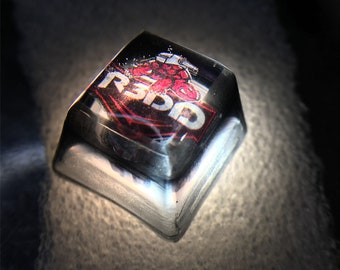 Pode incluir: Uma tecla de plástico transparente com um logotipo vermelho e branco que diz "Redd" e uma imagem de desenho animado de uma pessoa em um terno vermelho.